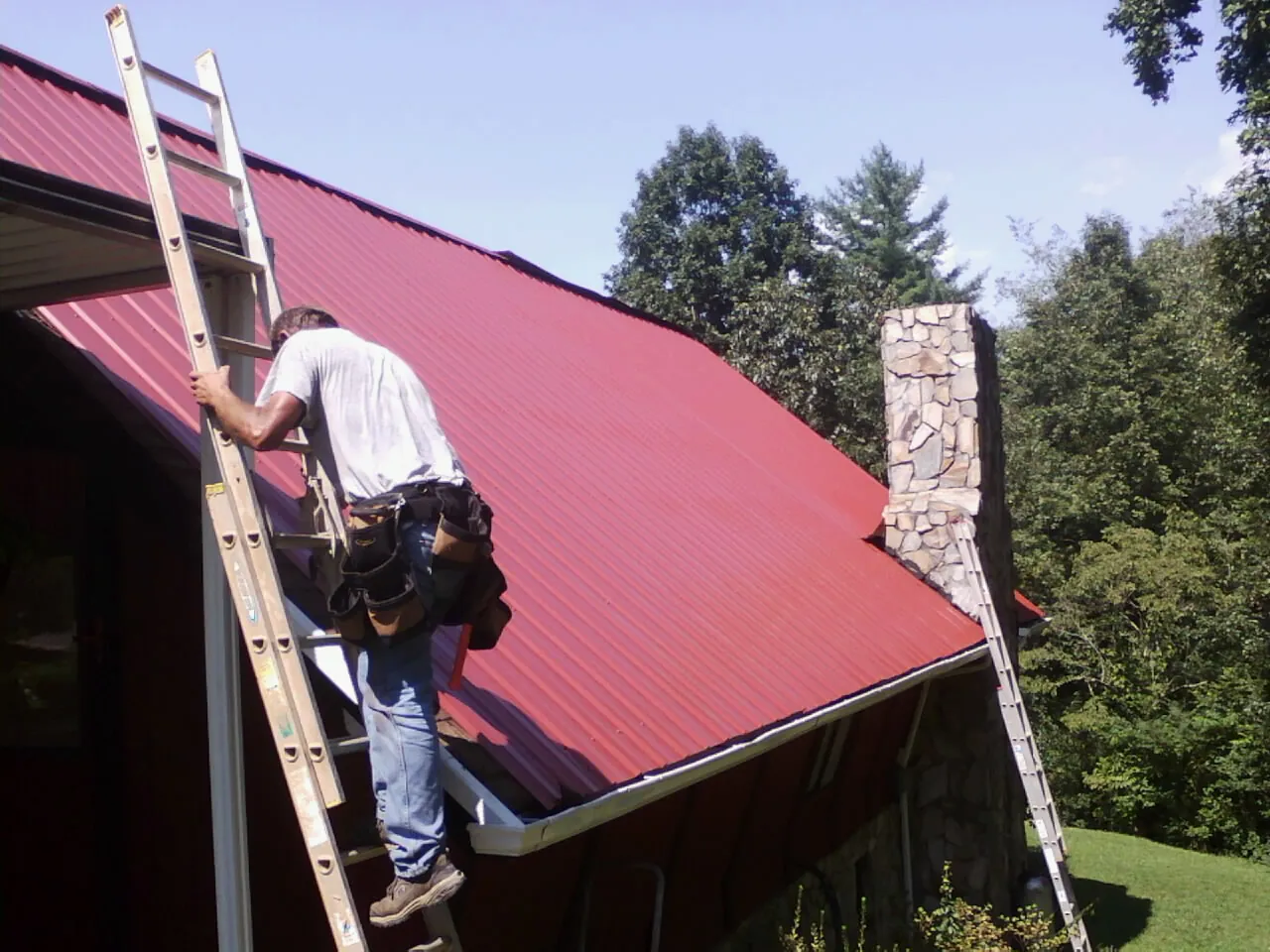 Roofer on a ladder beside a red metal roof and stone chimney — Soffit & Fascia Repair by Roofing Brothers in Crumpler